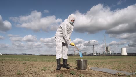 Soil-sampling-after-ecological-disaster,-scientist-digging-field-ground-with-factory-in-background