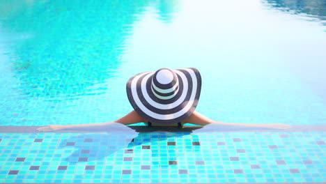 Back-to-the-camera,-a-woman-in-a-floppy-sun-hat-relaxes-in-a-resort-swimming-pool