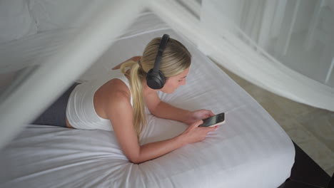 Woman-listening-music-on-wireless-headphones-in-bed-while-scrolling-on-smartphone