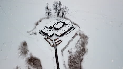 Snow-covered-wooden-buildings-surrounded-by-white-frozen-lake-while-snowing,-aerial-drone-view