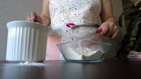 Latin-woman-wearing-an-apron-preparing-baking-a-cake-sieving-the-flour-using-a-metal-strainer-and-a-spoon-to-serve-it