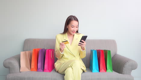 A-young-woman-in-a-pastel-business-suit-sits-among-colorful-shopping-bags-while-holding-a-smartphone-in-one-hand-and-a-credit-card-in-the-other