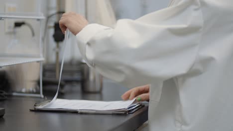White-coat-female-scientist-in-lab-reading-through-research-notes-on-a-clipboard