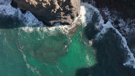 Aerial-shot-flying-over-turquoise-waves-crashing-against-rugged-cliffs-on-a-sunny-day