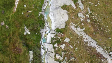 top-down-aerial-view-of-small-mountain-river-flowing-between-rocks-and-grass-with-a-hiking-path,-switzerland-hiking-paradise
