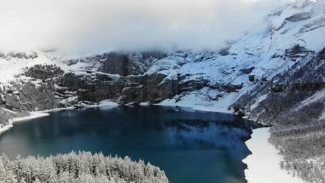 Luftüberführung-über-Die-Schneebedeckten-Bäume-Am-Ufer-Des-Oeschinensees-In-Kandersteg,-Schweiz-An-Einem-Wintertag-Mit-In-Wolken-Verborgenen-Gipfeln