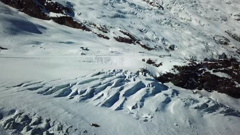 Blauer-Gletscher-Mit-Spalten-In-Den-Schweizer-Alpen,-Skiinfrastrukturen-Im-Hintergrund