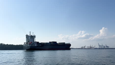 Container-Frachtschiff-Gefilmt-Von-Der-Seite,-Die-An-Einem-Sonnigen-Tag-Mit-Blauem-Himmel-Und-Ruhigem-Wasser-Im-Hafen-Ankommt