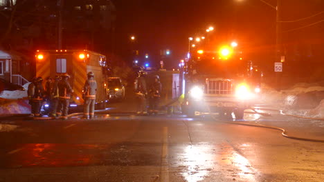 Group-of-firefighters-stand-on-the-street-in-front-of-fire-trucks-with-flashing-lights-on-a-cold-winter-night-in-the-city-of-Toronto,-Canada