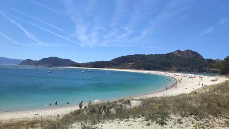 people-enjoying-afternoon-on-the-beach-with-sand-dune,-anchored-boats-and-traces-of-planes-in-the-sunny-sky,-panoramic-shot-bloueado,-Cíes-Islands,-Pontevedra,-Galicia,-Spain