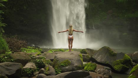 Woman-in-white-bikini-raising-arms-greeting-powerful-stream-of-waterfall,-Nungnung