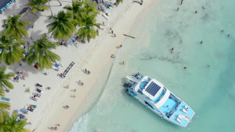 Local-tourists-walk-along-a-white-sand-beach,-a-luxury-party-yacht-anchored-on-the-shore,-aerial-top-down
