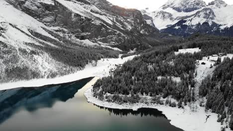 Aerial-flyover-over-shores-of-lake-Oeschinensee-in-Kandersteg,-Swtizerland-on-a-cold-winter-day-with-snow-covered-trees