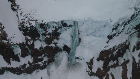 Water-Flowing-from-Waterfall-on-Icy-Snow-Covered-Iceland-Cliff---Aerial