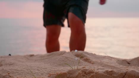 Asiatischer-Junger-Mann,-Der-Bei-Sonnenuntergang-Mit-Fokussiertem-Strandsand-Auf-Die-Kamera-Zugeht