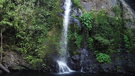 Drone-aerial-moving-backwards-a-tropical-green-waterfall-in-rainforest