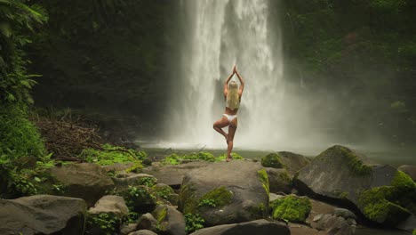 Mujer-Rubia-En-Equilibrio-De-Pose-De-Yoga-De-árbol-Con-Poderosa-Cascada-Nungnung