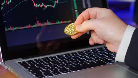 Geschäftsmann-Dreht-Gold-Bitcoin-In-Seinen-Fingern-Um,-Während-Er-Sich-Die-Börsencharts-Und-Preise-Auf-Einem-Laptop-Ansieht