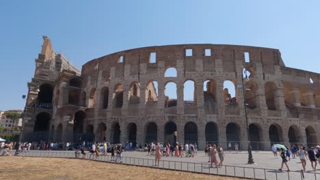 Establishing-shot,-iconic-Colosseum-monument,-group-tourists-walking-outside-Landmark,-Panning-Shot