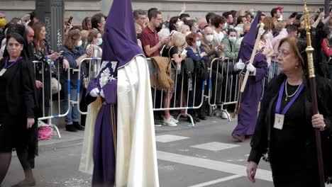 Nazarene-on-purple-tunic-and-hoods-and-Manolas-on-black-dresses-with-mantilla-carrying-candles-and-marching-between-crowds-during-Good-Friday,-Holy-Week,-in-streets-of-Madrid,-Spain