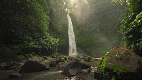 Woman-practicing-tree-pose-on-boulder-at-magnificent-Nungnung-waterfall-with-magical-sunlight