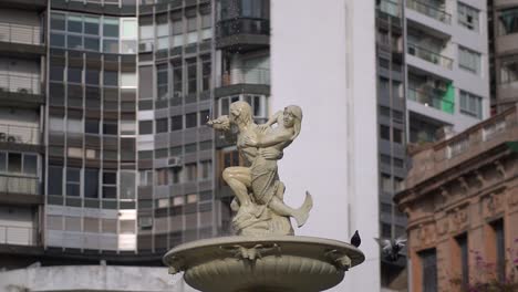 Slow-motion,-handheld-wide-view-of-Utopias-Fountain-in-Rosario,-Argentina,-against-a-backdrop-of-buildings
