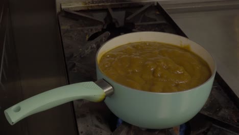 gravy-pot-in-light-teal-color-over-commercial-gas-heated-stove-next-to-a-stainless-steel-grill-zoom-out-surrounded-by-aluminum-counter-tops-with-semi-bubbly-gravy-topping-made-of-beef-animal-fat