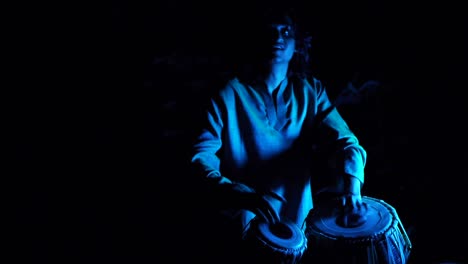 Indian-man-drumming-ecstatically-on-dual-tabla-setup-in-outdoor-location-with-hard-blue-key-light,-filmed-handheld-as-medium-shot