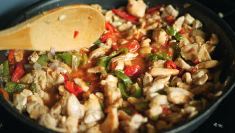 Close-up-of-wooden-spoon-mixing-chicken-fajitas-ingredients-cooking-in-pan