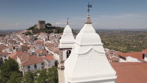 Antena-Dando-Vueltas-Alrededor-De-Los-Campanarios-De-La-Iglesia-De-Santa-Maria-Da-Devesa-Y-El-Antiguo-Castillo-En-El-Fondo,-Castelo-De-Vide-En-Portugal