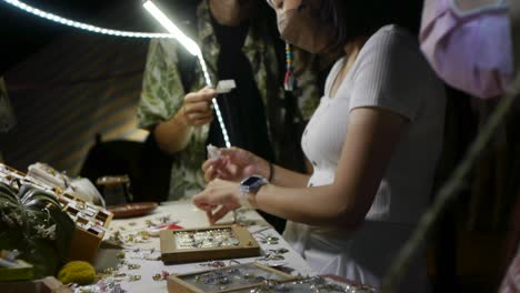 Asian-woman-wearing-face-mask-while-browsing-jewelry-at-outdoor-night-market,-filmed-handheld-with-slow-downward-tilt