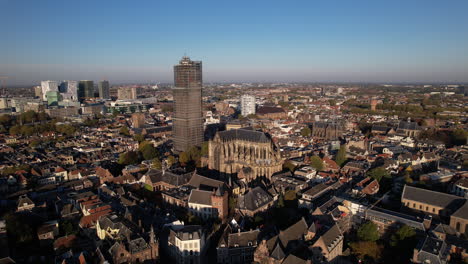 Aerial-sideways-pan-around-De-Dom-medieval-cathedral-tower-in-scaffolding-in-Dutch-city-centre-of-Utrecht-towering-over-the-cityscape-against-a-blue-sky-sunrise-and-orange-glow-on-the-horizon
