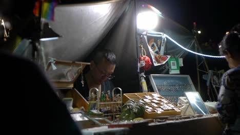 Asian-man-working-as-jewelry-vendor-at-outdoor-night-market,-filmed-in-handheld-style