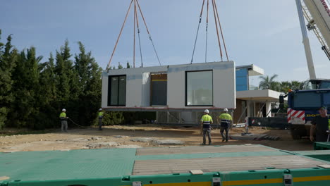 Over-truck-bed-to-workers-guiding-modern-modular-home-onto-foundation-with-crane-lift