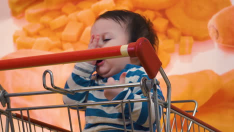 Tired-baby-hitting-himself-in-the-face-slightly-and-whining-while-waiting-for-family-inside-store-food-shop-grocery-sitting-in-shopping-cart