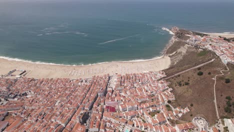 Aerial-orbiting-above-Nazare-village-and-white-sandy-beach-on-summer-day,-Portugal