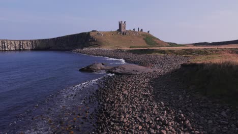 Slow-motion-drone-shot-flying-along-the-coastline-towards-Dunstanburgh-Castle,-Northumberland,-UK