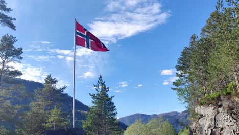 Norwegian-flag-waving-in-the-wind-surrounded-by-green-trees-and-with-blue-sky-background---Handheld-static-clip