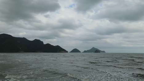 Low-altitude-reverse-aerial-footage-revealing-the-rushing-waves,-islands-in-the-distance,-mountains-silhouetting,-grays-clouds