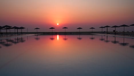 Perfect-red-sunset-view-from-tropical-hotel-resort-lounge-with-infinity-pool,-beach-umbrellas-and-their-reflection