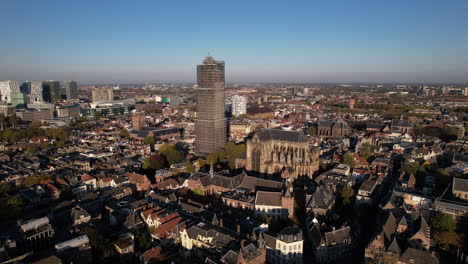 Sideways-aerial-view-of-De-Dom-medieval-cathedral-tower-in-scaffolding-in-Dutch-city-center-of-Utrecht-towering-over-the-cityscape-against-a-blue-sky-sunrise-and-orange-glow-on-the-horizon