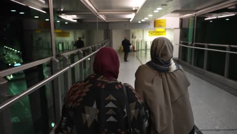 POV-Walking-Behind-Women-Wearing-Hijab-Making-Their-Way-Through-To-Departures-Terminal-T3-At-Heathrow-Airport,-Crossing-Over-Gangway-Bridge