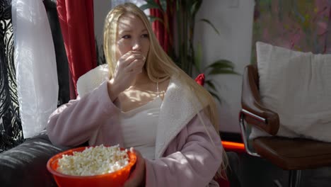Beautiful-blonde-white-caucasian-girl-women-sitting-on-couch-in-front-of-windows-in-pink-bath-robe-resting-relaxing-at-home-eating-popcorn-from-large-red-bowl-and-laughing-smiling-at-comedy-or-movie