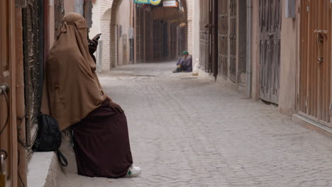 Marrakesh-woman-wearing-Burqa-talking-on-mobile-phone-and-homeless-man-sitting-in-narrow-Marrakech-alleyway