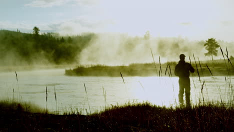 Cinematic-silhouette-shot-of-a-man-with-fishing-rod-in-his-hand-at-the-shore-of-river-illuminated-by-shiny-sunlight-and-smoke,-POV