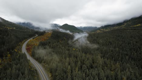 Valle-De-Montaña-Neblinoso-Aéreo-En-Movimiento-Con-Carretera-Con-Curvas,-Por-Encima-De-Las-Nubes-A-Través-Del-Bosque-De-Otoño