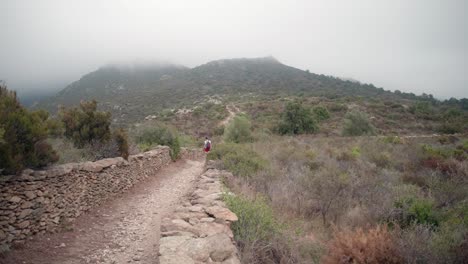 Female-Hiker-Walk-On-Dirt-Track-Towards-Foggy-Mountain-Near-Roses,-Catalonia,-Spain