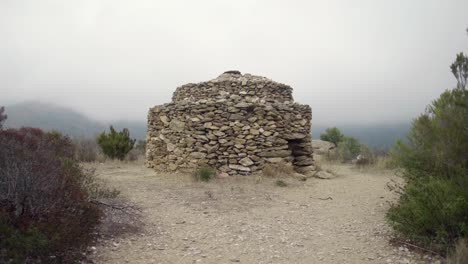 Antigua-Cabaña-De-Piedra-Cerca-Del-Sendero-Megalítico-Con-Una-Montaña-Nublada-En-El-Fondo-Cerca-De-Rosas,-Cataluña,-España