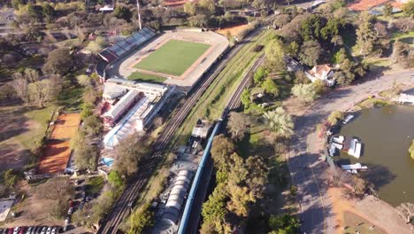 Blue-train-running-and-crossing-Palermo-area-with-ponds-and-Rosedal-gardens-in-Buenos-Aires,-football-field-in-background