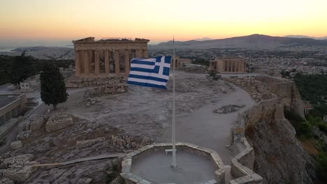 Cinematic-shot-of-the-flag-of-Greece,-Acropolis-city-of-Athens-parthenon,-Mount-Lycabettus,-Parliament-Building-and-residential-buildings,-sunset-in-Athens,-Greece